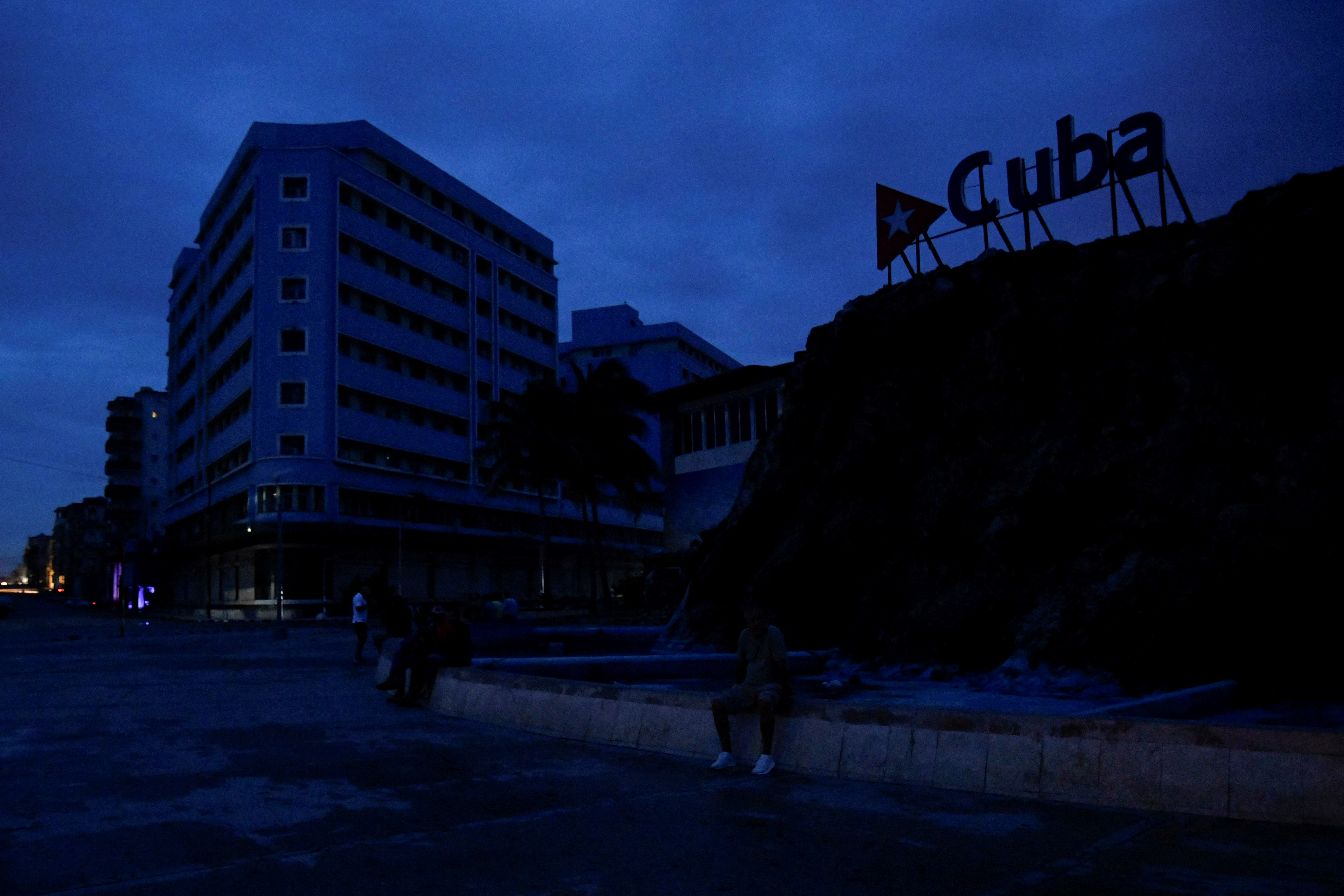 The silhouette of a sign that reads "Cuba" is seen against a blue sky, above a darkened street.