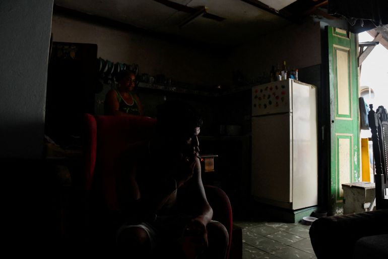 A person smokes a cigarette in his darkened home during a blackout, while sun pours in through an open door.